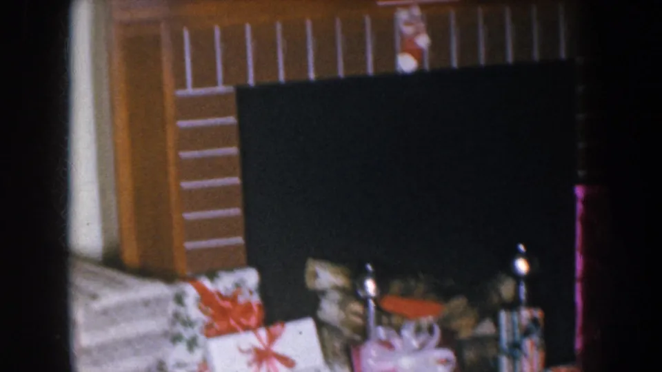 Wrapped Christmas gift boxes with red ribbons and bows piled near a dark fireplace surround and wooden mantel; from a mid-1950s Nebraska 8mm home movie.