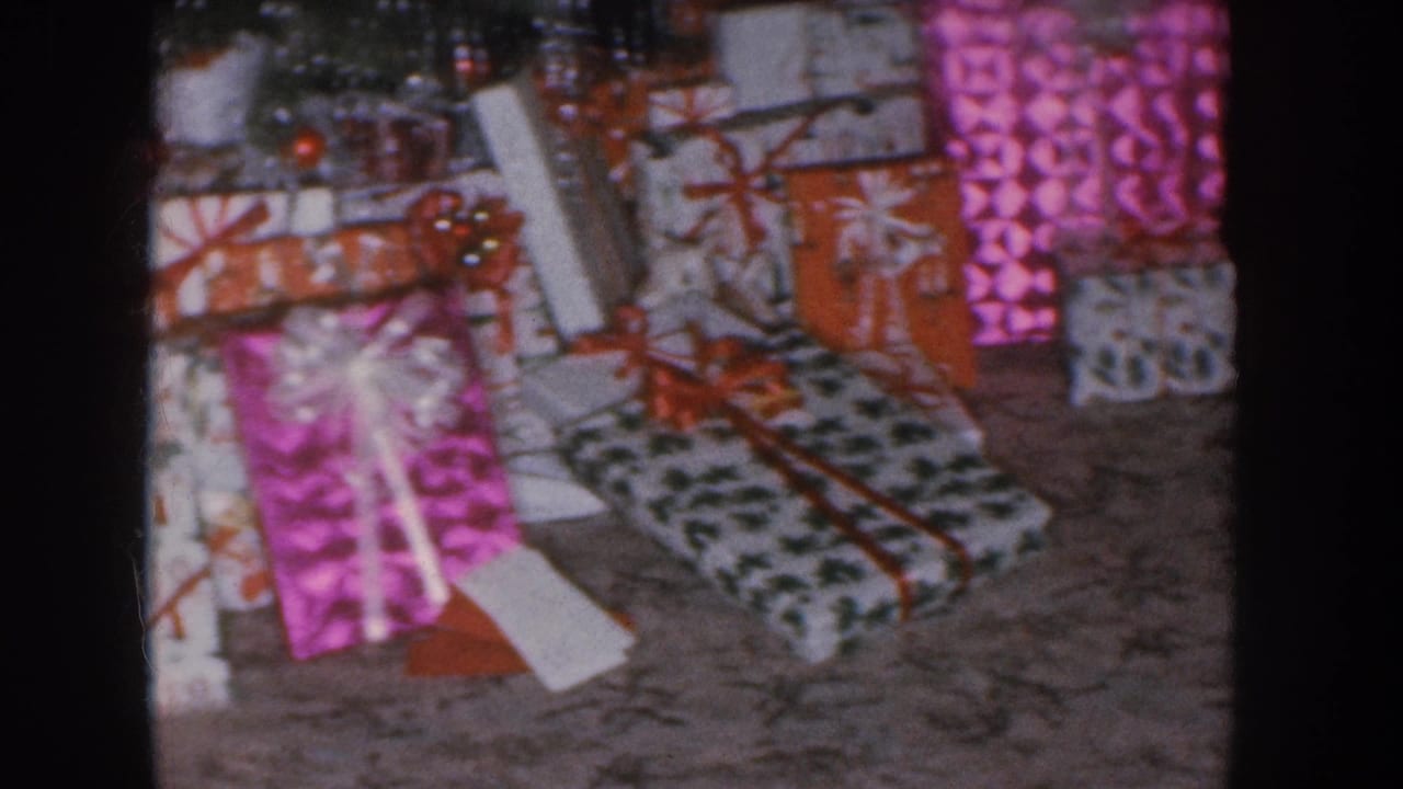 Mid-1950s home movie showing wrapped Christmas gifts in pink, orange, and black-patterned paper piled on a carpeted living room floor, Nebraska circa 1955.