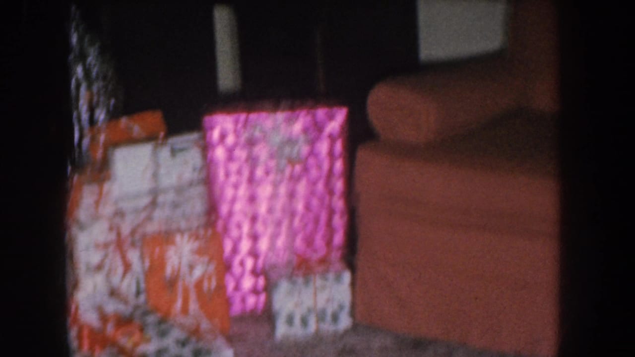 Close view of Christmas gifts wrapped in bright pink and orange patterned paper, with a salmon-colored sofa arm visible at upper right.