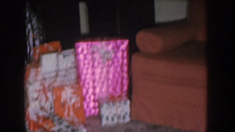 Circa 1955 home movie close-up of Christmas gifts in bright pink and orange wrapping paper; edge of a salmon upholstered sofa visible at upper right.