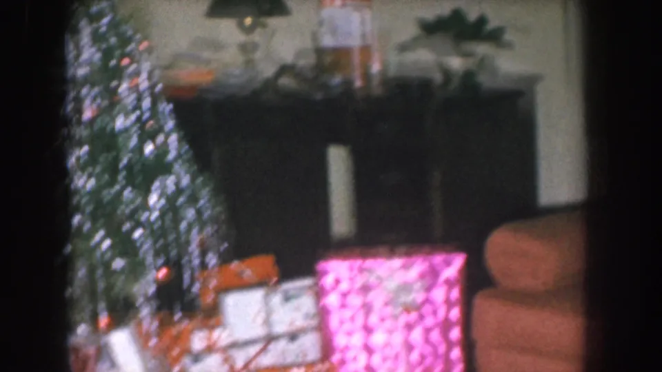 Mid-1950s living room with a lit Christmas tree at left, a dark wooden cabinet or fireplace surround with lamp behind, and wrapped pink gifts on the floor; salmon sofa visible.