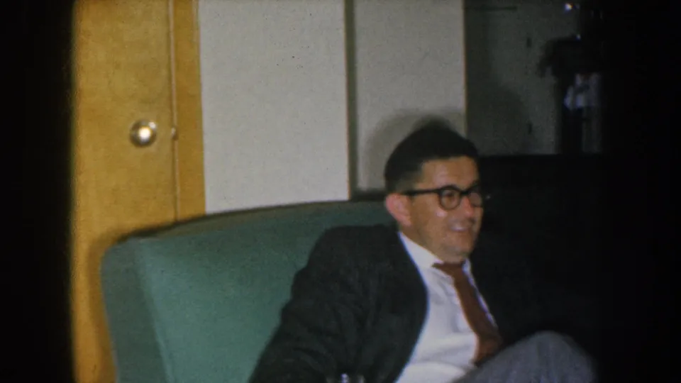 Mid-1950s home movie of an adult man wearing glasses and a dark tweed sport coat with tie, seated in a green armchair and smiling; yellow wall panel at left, circa 1955 Nebraska.