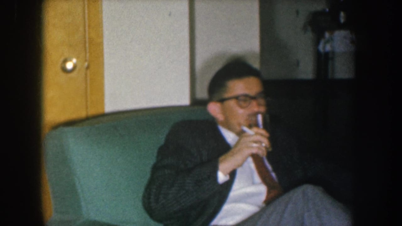Man in dark patterned suit and thick-rimmed glasses drinks from a glass on a green sofa; wooden door with brass knob and potted plant visible — Nebraska, USA, c. 1955, 8mm color.