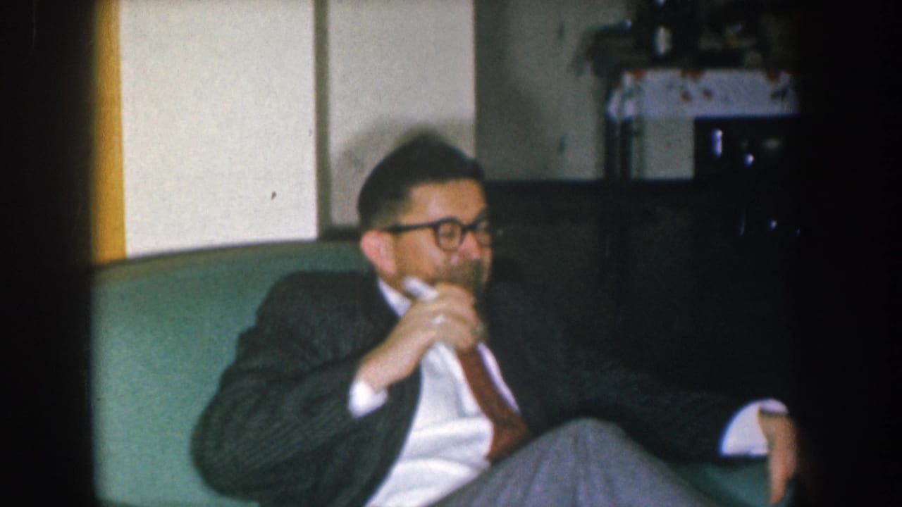 Man in dark suit and glasses with hands raised near his face on a green sofa; a second person's arm or sleeve is partially visible at the right edge.