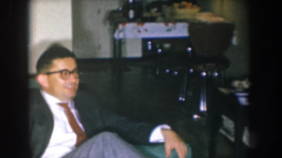 Man in dark patterned suit and glasses smiling toward camera on green sofa with lace-covered side table and snack bowl behind him — Nebraska, USA, c. 1955, 8mm color.