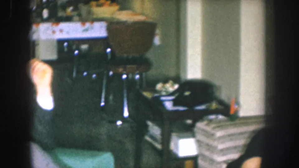 Wide living room shot: lace-covered side table with snack bowl and wicker basket, stacked magazines on the floor, and a second person's bare foot — Nebraska, USA, c. 1955, 8mm color.