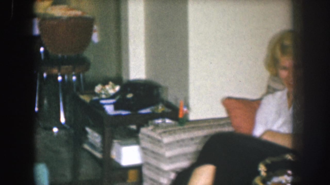 Blonde woman in a white blouse seated on a gray-striped sofa, looking right; a metallic side stand with stacked objects is visible in the background of what appears to be a 1950s domestic interior.
