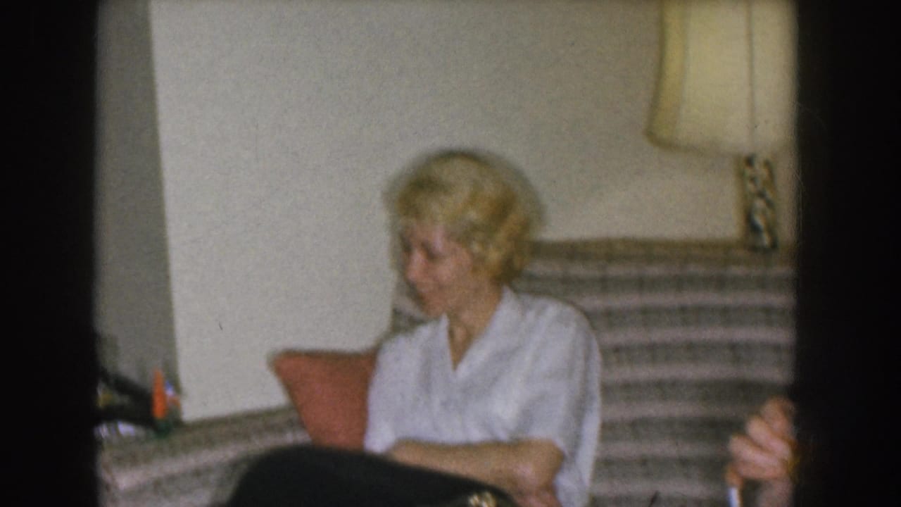 Blonde woman in white blouse on a striped sofa, facing slightly toward camera; table lamp with square shade visible at upper right.