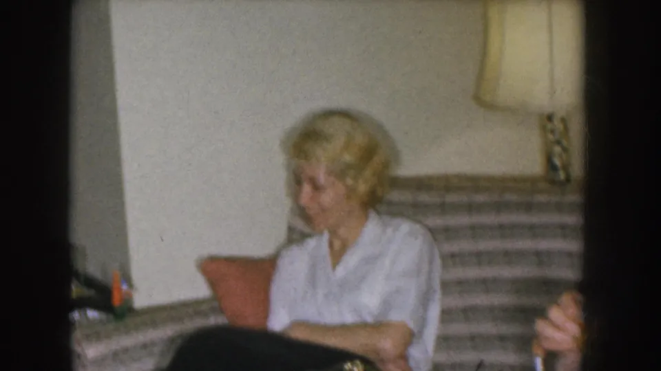 Blonde woman in a white blouse on a striped sofa facing toward camera; a table lamp with square shade is visible at upper right, consistent with a 1950s American domestic interior.