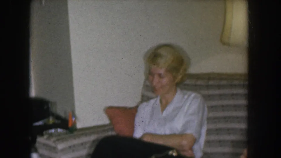 Blonde woman in a white blouse looking downward while seated on a striped sofa; small objects visible on a side surface at the left edge of frame, in what appears to be a 1950s domestic interior.