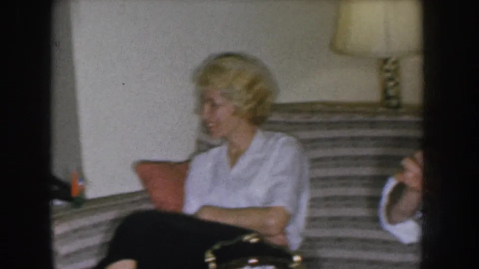 Blonde woman on a striped sofa in a 1950s domestic interior; a table lamp with chain pull is visible at upper right; a partial white object at the lower right edge may indicate a second person off-frame.