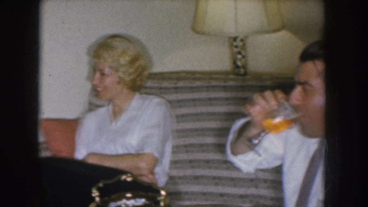 1955 Nebraska home movie — blonde woman in white blouse beside a man drinking from a glass; striped couch and table lamp visible in background.