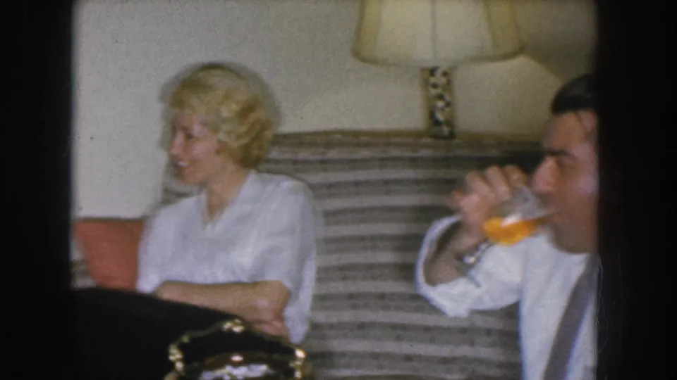 1955 Nebraska home movie — blonde woman in white blouse beside a man drinking from a glass; striped couch and table lamp visible in background.