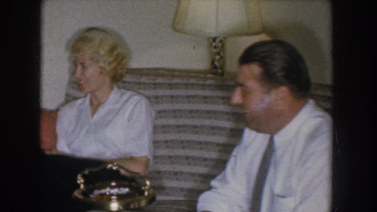 Blonde woman and man in white dress shirt and tie seated on striped couch in conversation; lamp and brass-rimmed object visible.