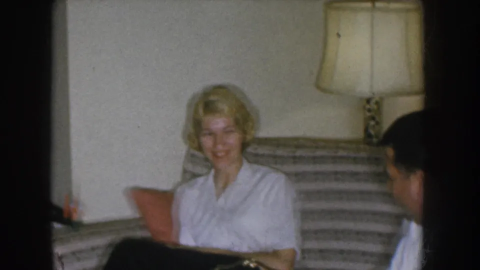 1955 Nebraska home movie — close view of a smiling blonde woman in a white blouse looking toward the camera during a social visit indoors.