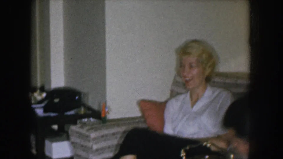 1955 Nebraska home movie — wider living room shot with a black object resembling a rotary telephone and a wicker basket in foreground; blonde woman and another figure on the striped couch.