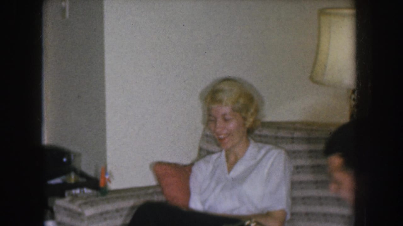 Blonde woman in white blouse and dark skirt, laughing, seated on a woven sofa in a 1955 Nebraska home interior.