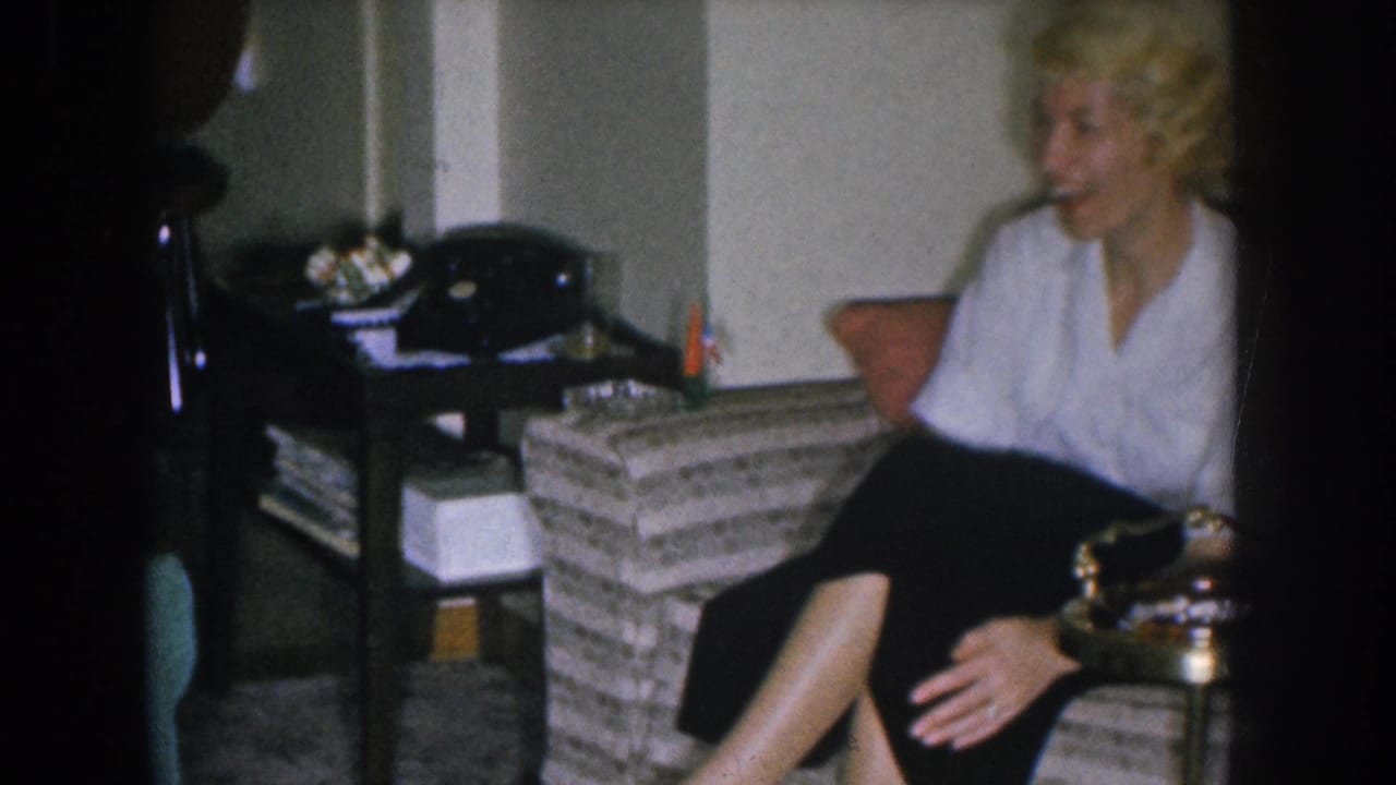 Blonde woman in white blouse and dark skirt seated, with a black rotary telephone and woven side table visible.