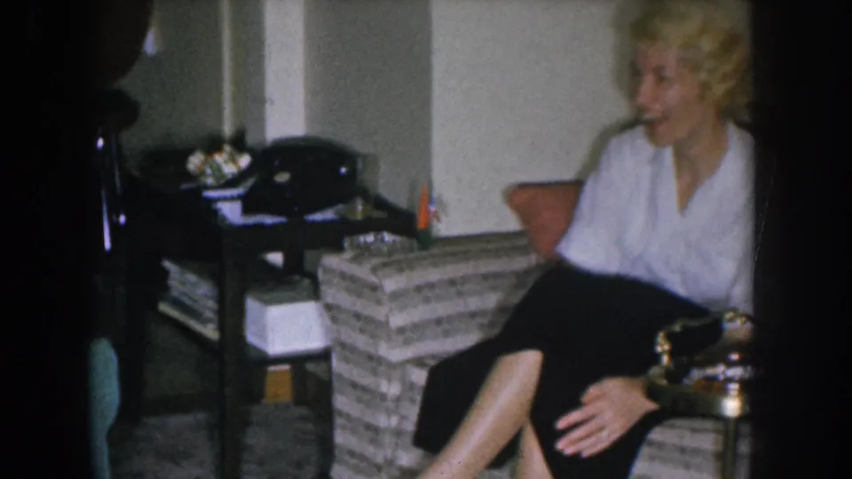 Blonde woman in white blouse and dark skirt mid-conversation in a 1955 mid-century American living room; rotary telephone and woven furniture visible.