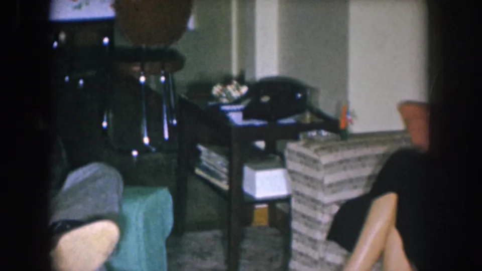 Living room interior circa 1955 with woven rattan side table, black rotary telephone, ashtray, and partially visible seated adults.