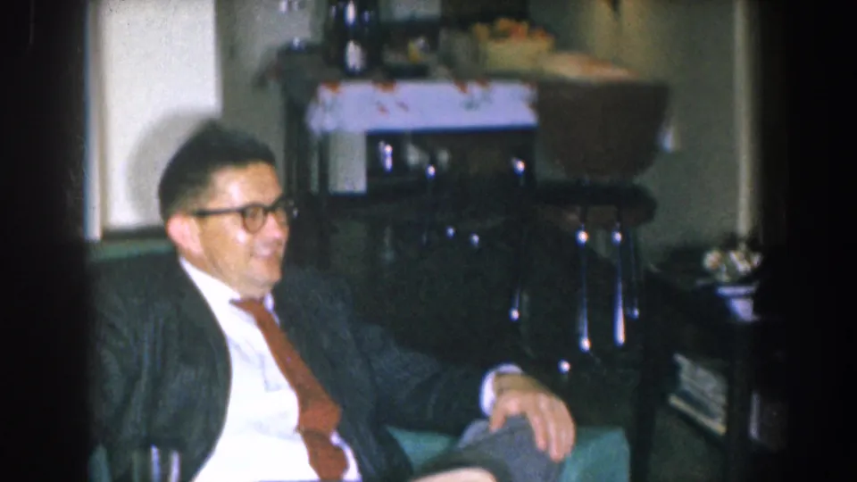 Man in dark suit with black-rimmed glasses and brown tie, smiling in a 1955 home interior; furnishings and objects visible behind him.