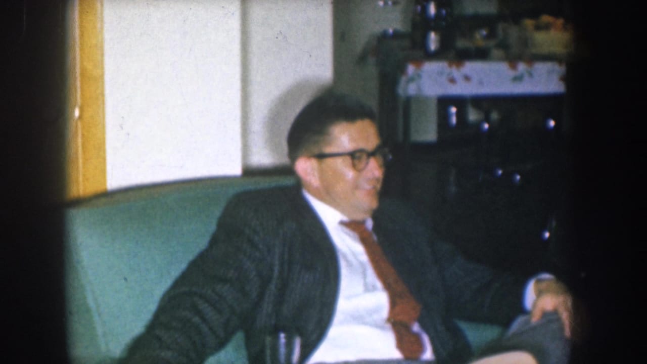 Mid-1950s Nebraska home movie — man in dark suit and thick-rimmed glasses smiling on teal couch during a casual social gathering.