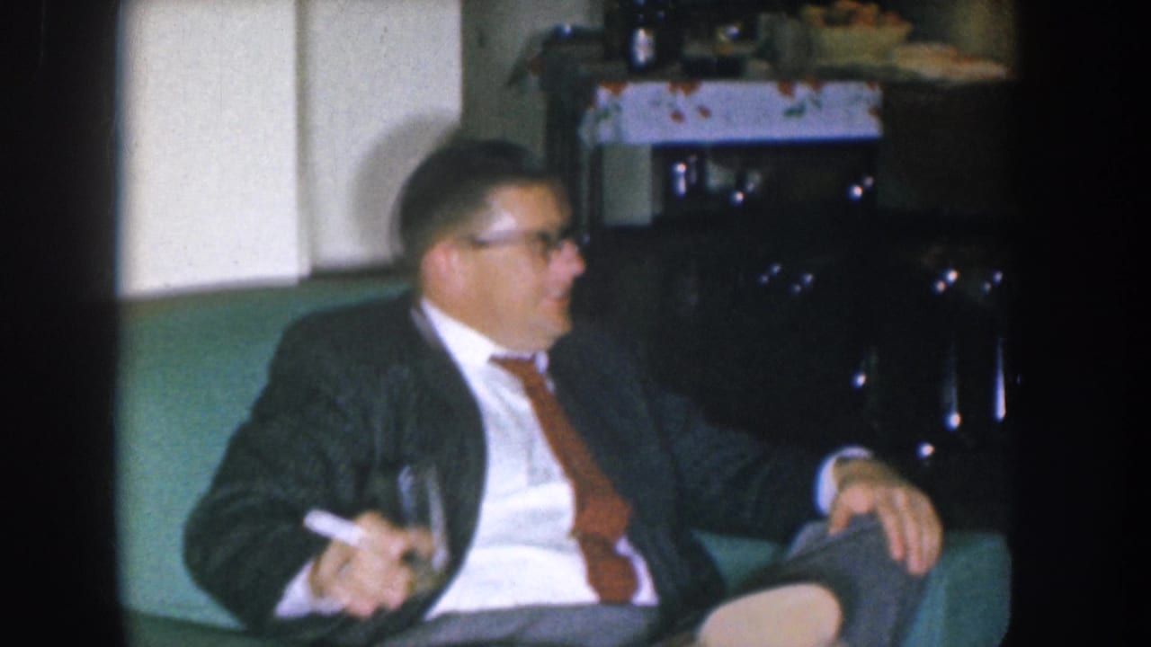 Man in dark suit holding cigarette and glass, seated on teal couch, looking off to his right.