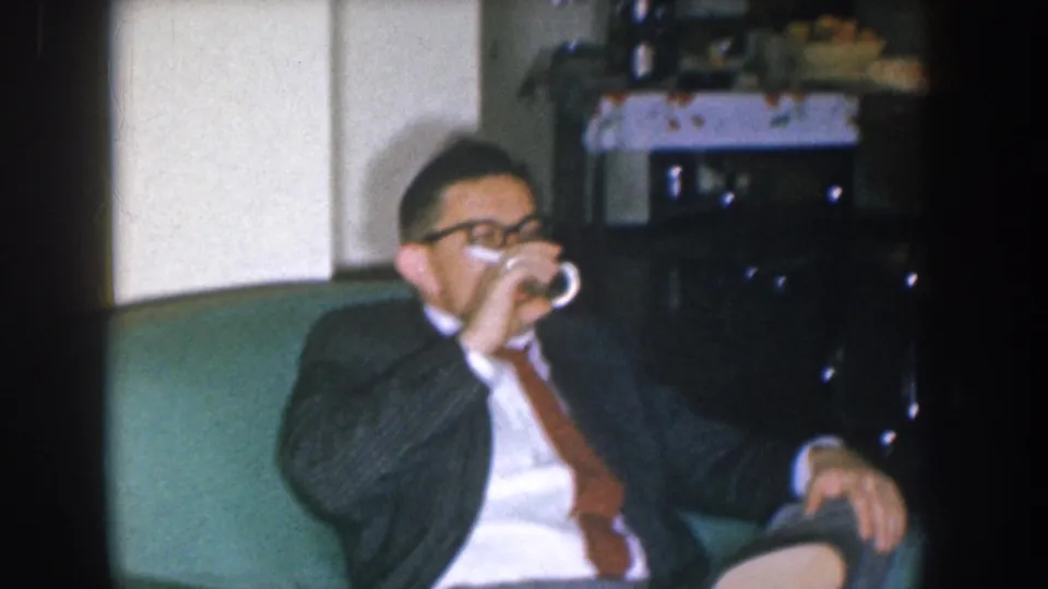 Mid-1950s Nebraska home movie — man in dark suit drinking from glass on teal couch, candid unscripted moment.