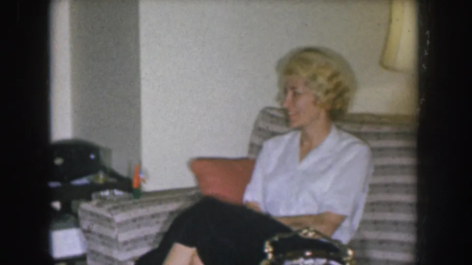 Mid-1950s Nebraska home movie — blonde woman in white blouse seated during a casual social gathering, domestic interior.
