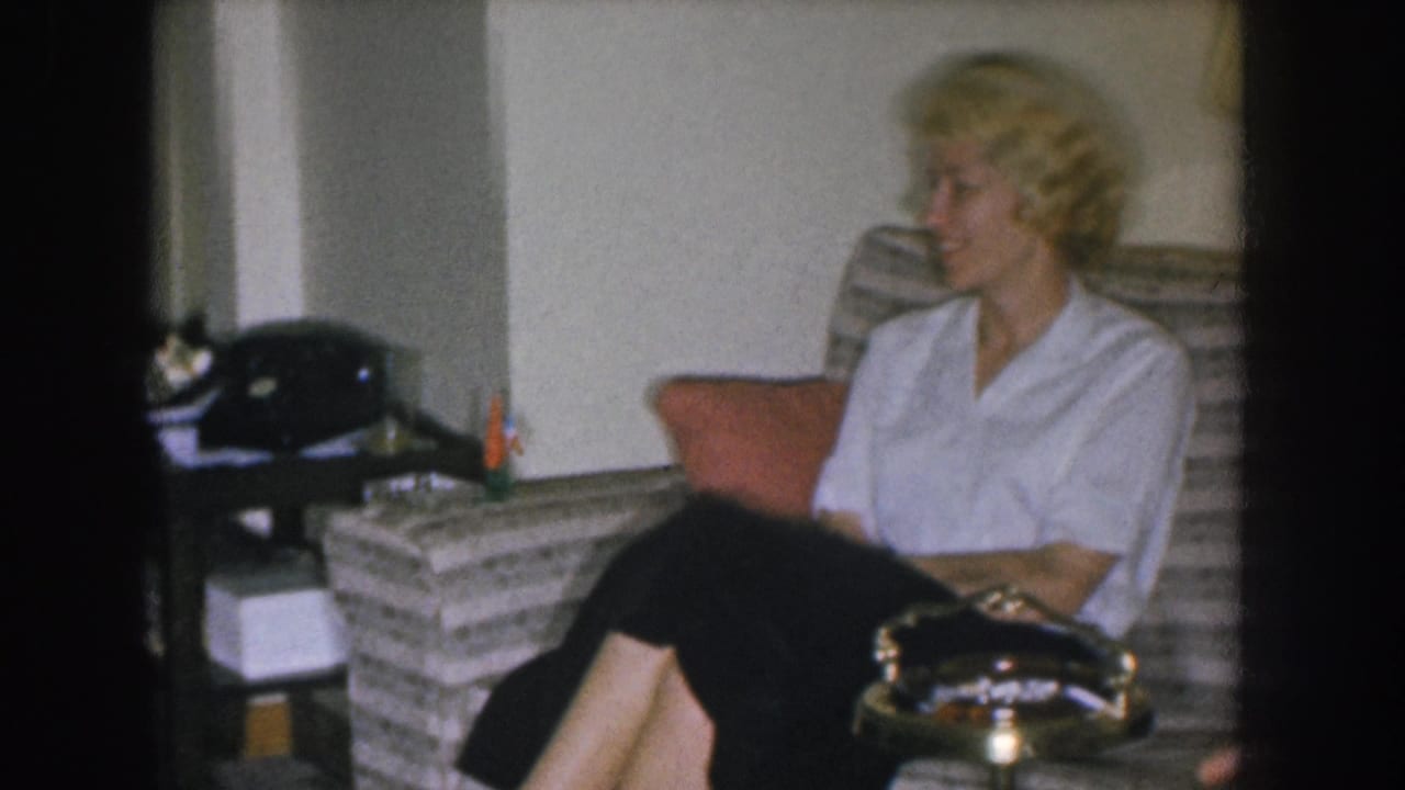 Blonde woman seated alone on a striped mid-century couch in what appears to be a 1955 Nebraska home interior; rotary telephone and brass ashtray visible.