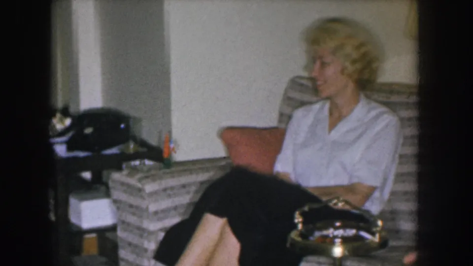 Blonde woman seated alone on a striped mid-century couch in what appears to be a 1955 Nebraska home interior; rotary telephone and brass ashtray visible.
