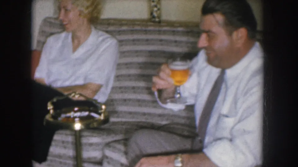 Couple on a mid-century striped couch circa 1955; man in white shirt and narrow grey tie holds a beer glass while woman smiles beside him.