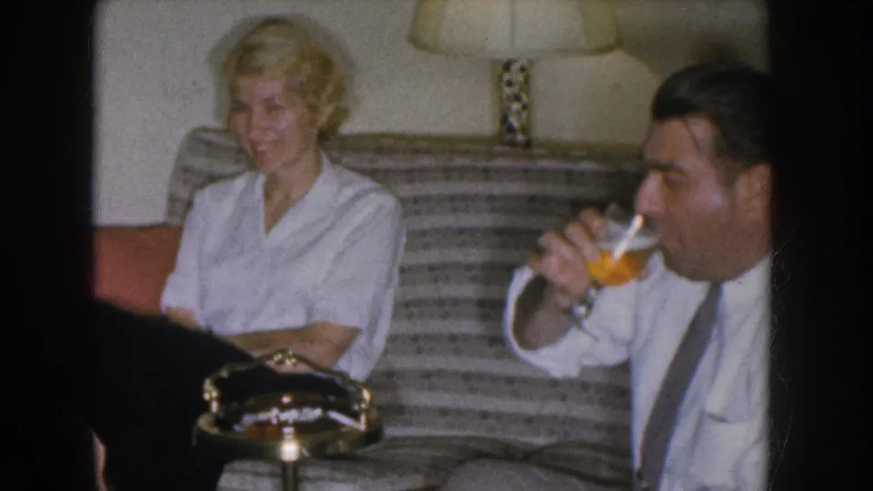 Couple in apparent 1955 Nebraska living room; woman smiles at man raising beer glass; mid-century floor lamp and striped upholstered couch visible behind them.