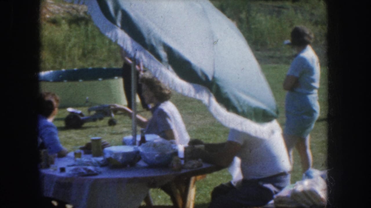 1955 Nebraska backyard family gathering, adults seated at a picnic table under a fringed umbrella, a woman standing nearby, 8mm home movie.