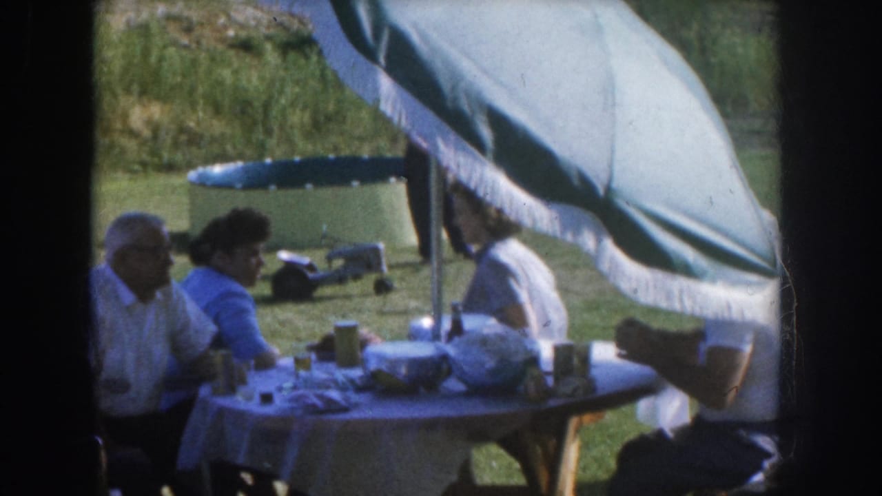 Several adults seated at an outdoor table under a fringed canopy; a teal inflatable pool is visible in the background lawn.