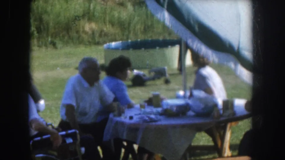 Nebraska backyard gathering, circa 1955, adults in close conversation at a cloth-covered outdoor table, large fringed umbrella overhead, inflatable pool in background.