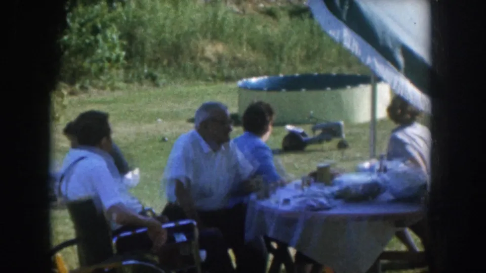 Mid-1950s Nebraska backyard gathering, adults at a picnic table, man in a dark cap and man in white shirt visible, fringed umbrella overhead, inflatable pool behind.