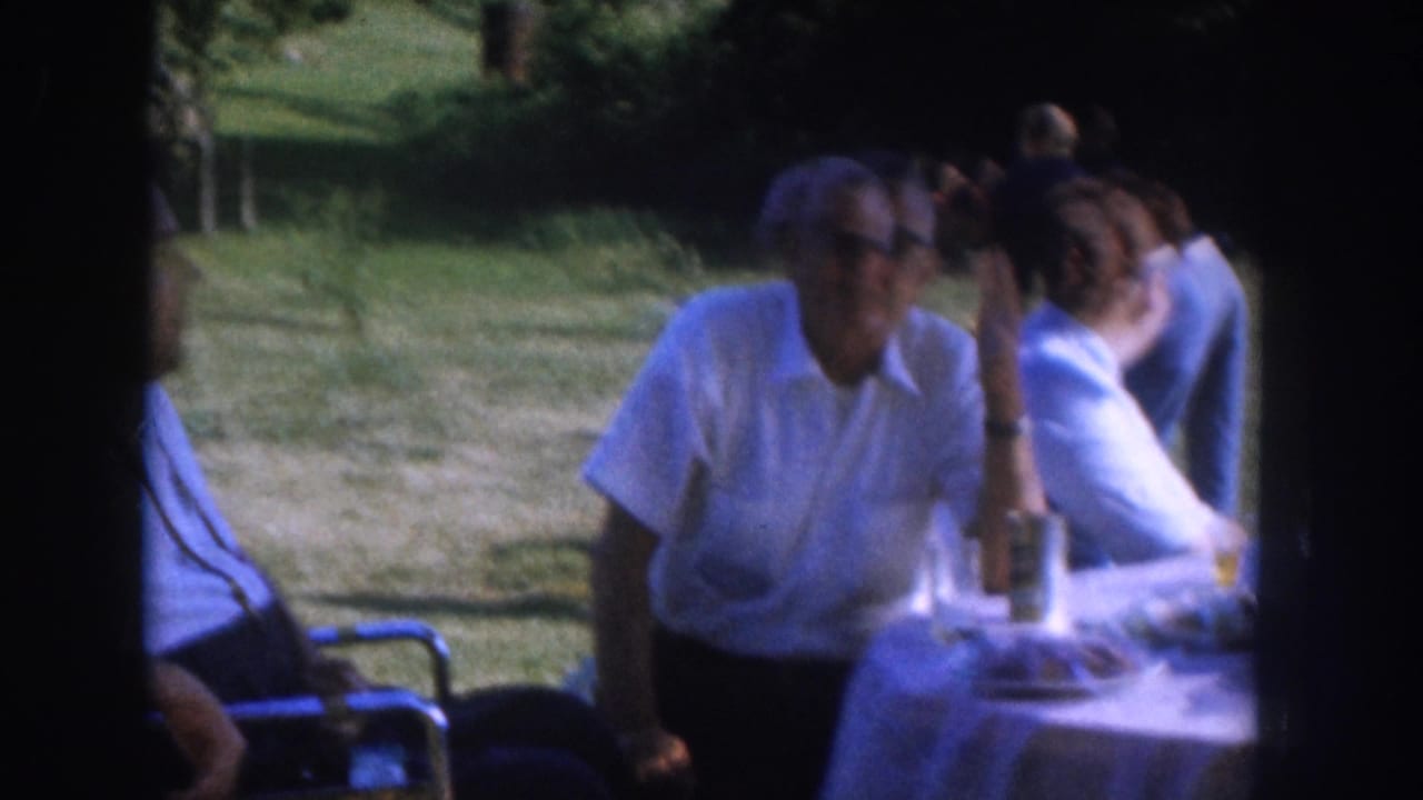 Outdoor gathering c.1955, Nebraska: older man with mid-century glasses at a white-clothed table, woman in light blue shirt behind him, wheelchair visible at left, green lawn and trees behind.