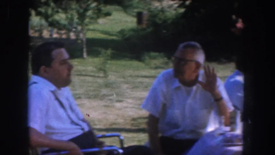 Two men in light shirts at a summer outdoor table c.1955; the right-hand figure gestures with a raised hand during conversation, a wristwatch visible; green lawn and trees behind.