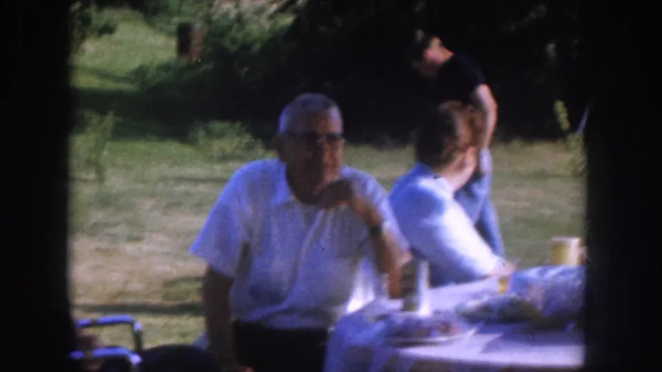 Older man with mid-century eyeglasses in white shirt at a white-clothed outdoor table, cups on the surface, second person partially visible at right, green lawn and trees behind, c.1955 Nebraska.