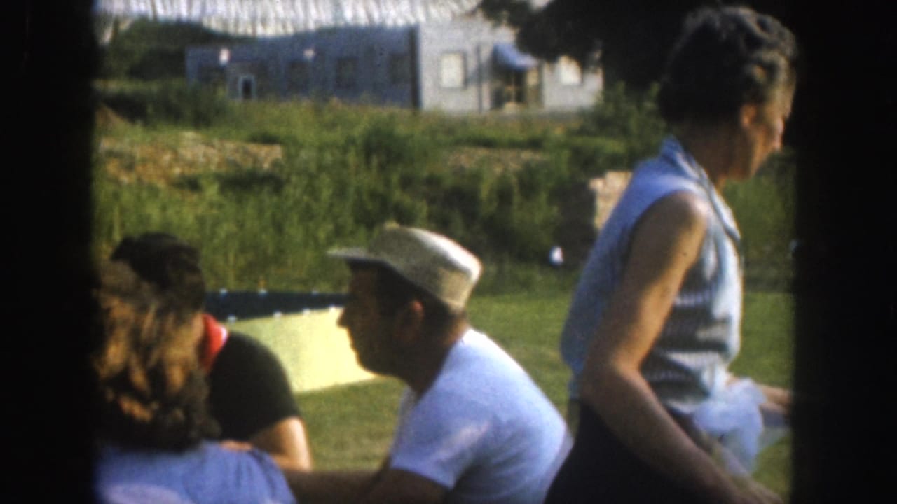 Mid-1950s outdoor gathering in Nebraska, USA: a standing woman in a sleeveless blue top and a seated man in a flat cap converse near a lush green lawn.