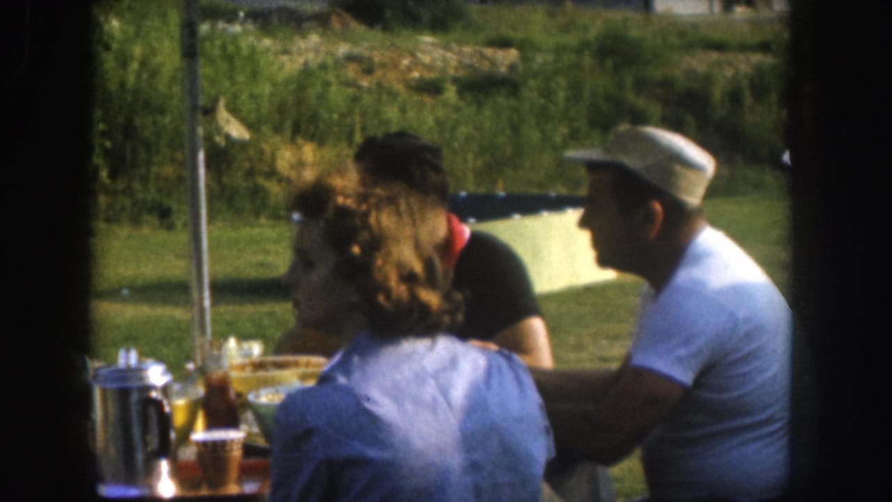 Two adults at an outdoor picnic table; a metal percolator and glasses of amber liquid are visible on the table.
