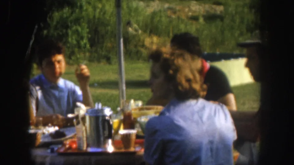 A candid 1950s outdoor picnic scene in Nebraska showing several adults around a table with plates, a metal percolator, and amber drinks on a bright summer day.