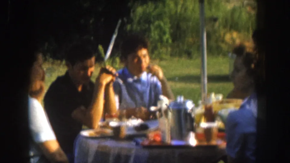 A dark-haired woman and a man in a white shirt sit at a 1950s outdoor picnic table in Nebraska with drinks, plates, and a red food item visible in the foreground.
