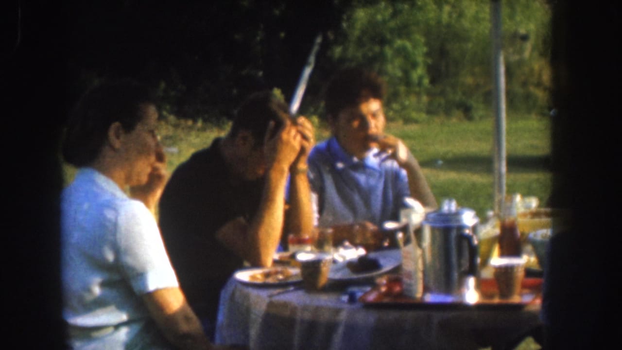 Three adults at a patterned cloth-covered outdoor table in what appears to be a Nebraska backyard, circa 1955, shot on 8mm film.