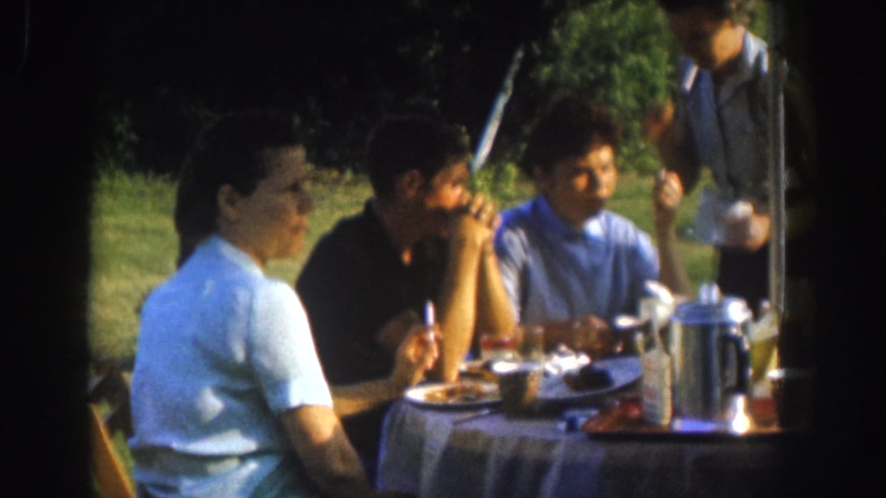 Woman turns toward camera at an outdoor table with two men; a fourth figure is partially visible standing at upper right.