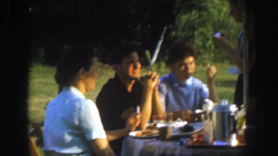 Mid-shot of three adults eating outdoors at a cloth-covered table, circa 1955; woman's back is toward the camera with a grassy yard and trees behind.