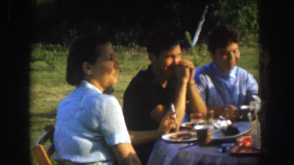 Closer framing of three adults sharing an outdoor meal in a grassy backyard, consistent with a Nebraska family gathering circa 1955, captured on 8mm.