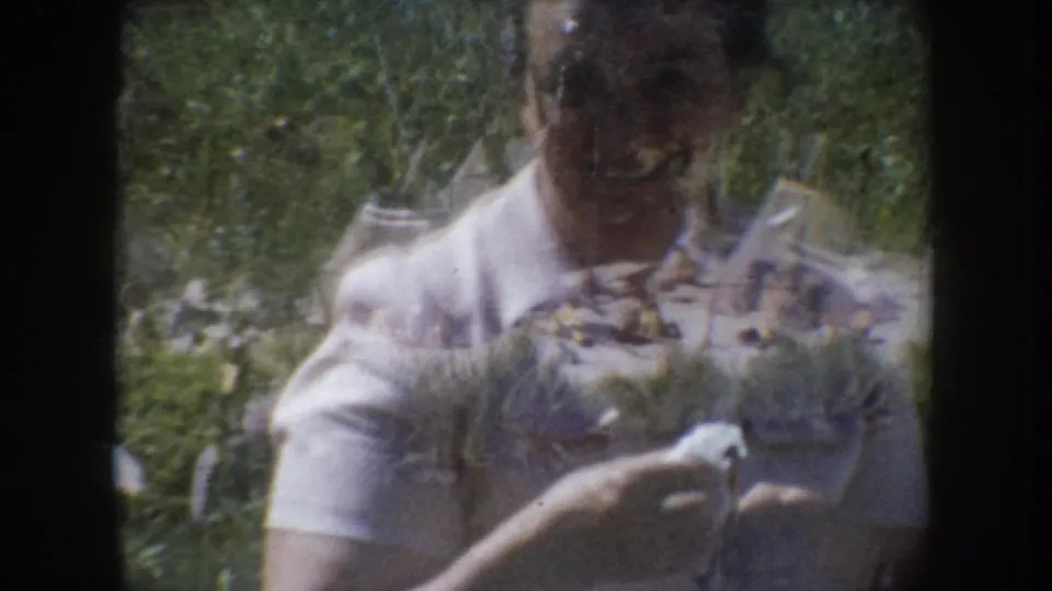 Woman in white collared shirt captured mid-motion outdoors, circa 1955 South Dakota; background begins to reveal a ground-level miniature display.