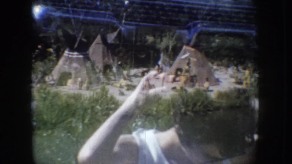 Roadside or tourist-site miniature village display with small model structures on grass, circa 1955 South Dakota; partial blue vehicle and woman's arm visible at frame edges.